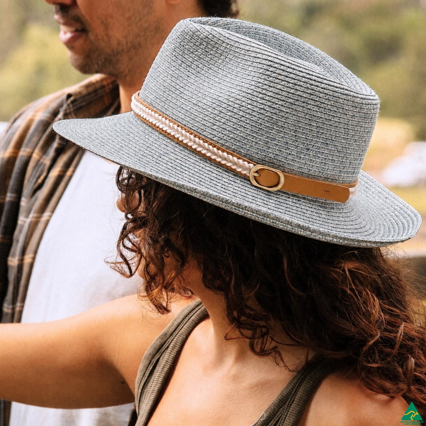 A woman wearing a light gray woven sun hat with a tan leather band and pearl-like trim, photographed outdoors with a man partially visible in the background.