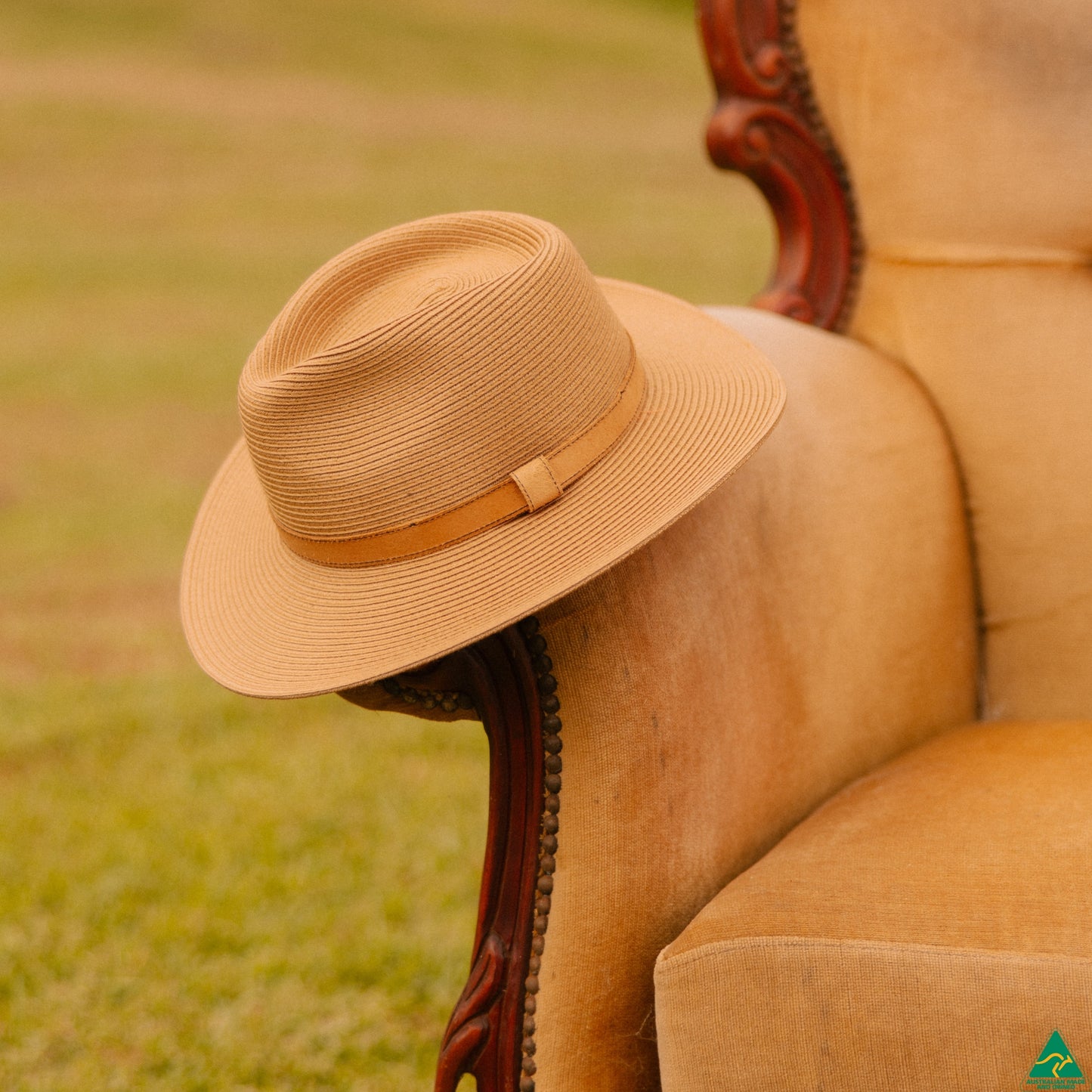 A beige woven fedora hat with a matching band rests on the arm of a vintage tan armchair, with a blurred grassy lawn in the background.
