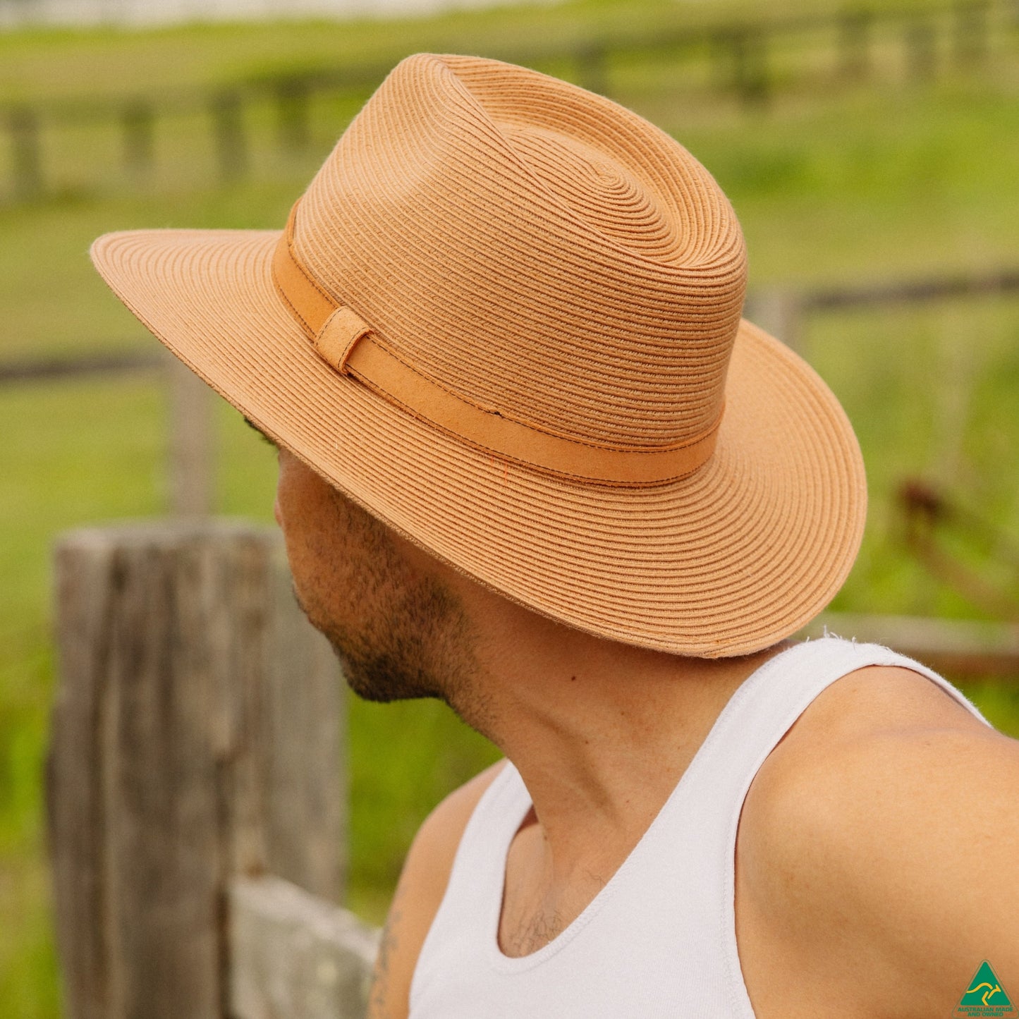 A man wearing a tan woven wide-brim hat with a matching band, photographed outdoors in a blurred green field setting.