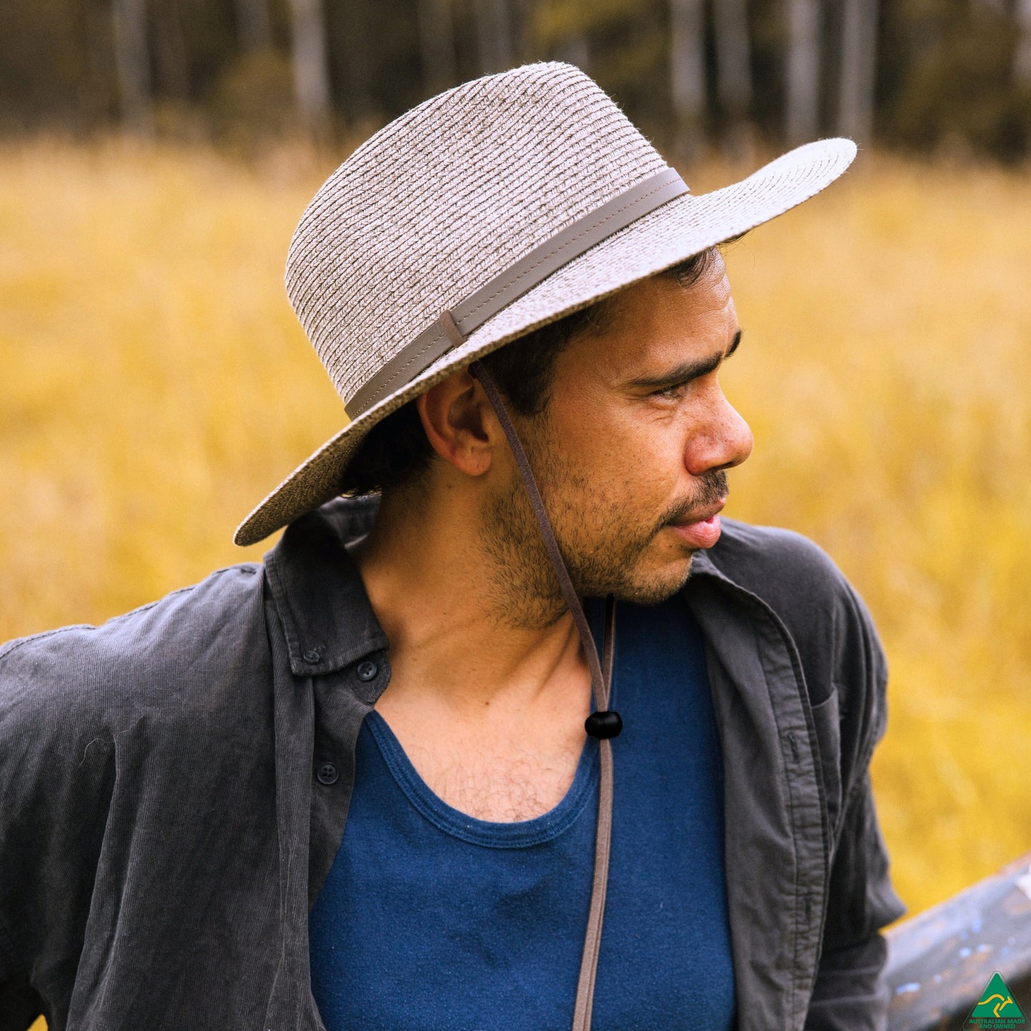 A man wearing a light gray woven straw hat with a dark band, a dark gray button-up shirt, and a blue tank top, standing in a field of dry yellow grass.